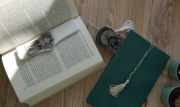 Handmade fabric bookmarks featuring animal illustrations of an elephant and Cat, placed on an open book and a closed green notebook.